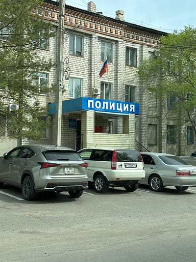 The image depicts the Police station in Raychikhinsk, Russia. The building is made of white brick and has a blue sign with the word "ПОЛИЦИЯ" (Police) written vertically in Russian. A Russian flag is mounted on the building. Three cars are parked in front of the station. The first car is a gray Lexus with the license plate "E 080 XM 28". The second car is a white Honda with the license plate "K 430 XM 28". The third car is a silver Toyota with the license plate "B 069 KX 28". Trees are present on either side of the building. Overhead power lines run across the top of the building. The sky is overcast.