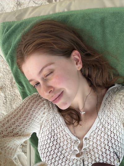 A young woman with red hair and freckles is lying on a green towel on a sandy beach. She is wearing a white knitted top and several necklaces, one of which has a pendant shaped like the letter "B." She is winking and smiling at the camera. The scene appears to be a casual vacation setting, possibly on a beach in Ao Cho, Thailand.