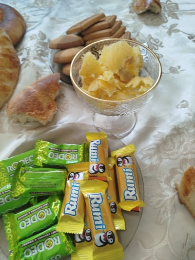 The image depicts a spread of food items on a tablecloth with a floral pattern. In the foreground, there are several wrapped candies, some in green packaging labeled "RODEO" and others in yellow packaging labeled "Rumi" with cartoon penguin characters. A plate with more of these candies is visible behind the initial layer. To the left, there are pieces of bread, one appearing to be a larger loaf and another a smaller, torn piece with a golden-brown crust. In the background, a glass bowl contains a yellow, shredded substance, possibly fruit or cheese. There are also several flat, rectangular cookies stacked on a plate. The setting appears to be a domestic environment, potentially a table laid out for a meal or snack. No people are visible in the image. The location is identified as Жаңы-Айыл, Kyrgyzstan.