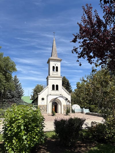 The image displays the Capilla Nuestra Señora de Luján in El Bolsón, Argentina, under a bright blue sky with wispy cirrus clouds. The central subject is a white chapel with a stone-clad foundation base. It features a prominent steeple topped with a shingled roof and a dark cross finial. The steeple's bell tower contains arched openings. The chapel's main facade includes an arched entrance framed by a dark metal trellis, with a small round window to its left and two lower rectangular windows positioned symmetrically above the stone base.
In the foreground, dense green foliage, including shrubs and a large leafy bush, frames the paved plaza in front of the chapel. The plaza surface is constructed from reddish-brown pavers. To the immediate right of the chapel entrance, a curved white stone bench or low wall is visible, with a dark metal gate positioned behind it. Further right, a tree with dark red-purple leaves partially obscures the view. The background consists of a dense arrangement of various green trees, and a building with a distinct green roof is partially visible behind the chapel to the left. The scene is illuminated by strong daylight, casting shadows. No individuals are present, and no specific actions or interactions are depicted.