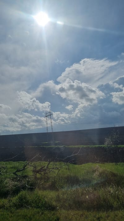 The scene, identified as being located in or near Suvorovo, Bulgaria, depicts an expansive outdoor landscape under bright, direct sunlight from the upper left, causing a distinct lens flare. The sky above is partly cloudy, featuring scattered white and grey cumulus formations against a blue background. Horizontally traversing the mid-ground is a dark, elevated landmass or embankment. A metal lattice transmission tower (pylon) stands prominently atop this landmass, slightly left of center, supporting multiple overhead power lines that extend horizontally across the frame. Additional smaller pylons are faintly visible further to the left. The foreground consists of green vegetation, predominantly grass, with several bare, fallen tree branches or roots lying on the ground in the lower-left and central portions. A small tree or bush with light-colored foliage is present near the right edge of the frame, positioned against the dark landmass. No human subjects or active interactions are observed.