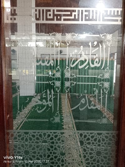 The image shows a view through a glass door with etched Arabic calligraphy on it. The door appears to be a doorway to a mosque, as the inscription on the door is in Arabic and there is a green prayer rug visible through the glass. The rug appears to be folded in half with part of a room visible behind it. The calligraphy on the door is in a white design with a black outline. The door is made of wood with a brown stain. The frame of the door has a geometric design. The scene is likely in a mosque in Kecamatan Gabuswetan, Indonesia. The image was taken on March 13, 2023, at 5:27 PM, with a Vivo Y19s smartphone.