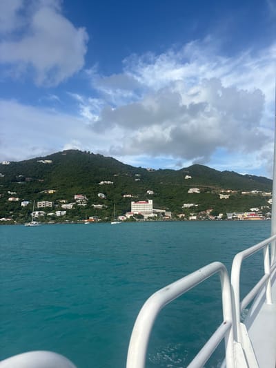 The image depicts a panoramic view of the coastline of Road Town, British Virgin Islands, as observed from the water. In the foreground, white railings of a moving vessel are visible on the right and bottom left, indicating the perspective is from a boat. The water is a clear, turquoise blue with minor surface ripples. In the midground, the verdant, steep hillsides of Road Town rise directly from the sea, covered in dense green vegetation. Numerous white and light-colored structures, primarily residential buildings, are scattered across the slopes. A prominent multi-story white building with a red roof, consistent with Maria's by the Sea Hotel, is centrally located on the shoreline. Several smaller boats, including at least one sailboat, are anchored in the bay between the viewing vessel and the shore. The sky above is a mix of deep blue and scattered white and grey clouds, with larger cloud formations towards the right. No individuals are discernible in the scene.