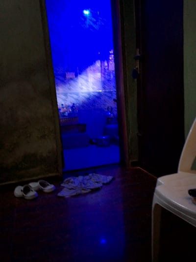 This image captures an interior scene in Nibo, Nigeria, viewed from a dimly lit room through an open doorway into an adjacent room illuminated intensely by blue light.
In the immediate foreground, a dark, possibly reddish-brown floor is visible. To the left of the doorway, a pair of white, closed-toe shoes is placed neatly on the floor. Adjacent to the shoes and slightly further into the room, a crumpled light-colored cloth object lies on the floor. On the right side of the foreground, the upper portion of a white plastic chair is partially visible, with a dark, small object resting on its seat. The wall to the left of the doorway is dark and appears to have a rough texture.
The doorway itself features a dark frame, and the dark-colored door is ajar to the right, revealing a latch mechanism. Beyond the doorway, the inner room is bathed in a bright, pervasive blue light, suggesting an artificial light source, possibly an LED fixture. Within this blue-lit room, objects are discernible but somewhat obscured by the light and atmospheric effects. These include what appears to be a white ceramic toilet or similar fixture on the far left, and various bottles and containers (possibly personal care products) on a surface or shelf. A metal pot or bowl is visible on the floor further into this room. The walls of this inner room appear to have a light-colored, possibly tiled or textured finish, reflecting the blue light. No individuals are visible in either room. The overall environment suggests an indoor domestic or commercial setting with distinct lighting choices.