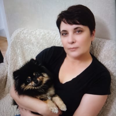 An adult woman with short, dark hair and light skin is sitting on a textured, light-colored sofa, holding a small black and tan dog. She is wearing a black V-neck t-shirt. The dog, which appears to be a Pomeranian, is nestled in her arms and facing the camera with its mouth slightly open. The background consists of a lightly textured wall. The scene appears to be indoors, possibly in a residential setting. The image quality is somewhat grainy, suggesting it may have been taken with a lower-resolution camera or under suboptimal lighting conditions. The location is specified as Chekhov-2, Russia.
