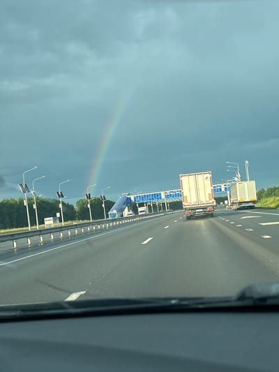 This is a photograph taken from a vehicle moving along a highway in Opytnoe Pole, Russia. The perspective is from inside the vehicle, looking forward and slightly to the left. The highway has multiple lanes marked with white lines. On the right side of the highway, the edge of the vehicle's interior is visible, suggesting the photo was taken with a phone.

The primary subjects are two trucks on the highway, one directly ahead of the other. Both are white with some markings. There are also overhead structures that appear to be part of a toll booth or gantry system, with blue panels and lighting. Along the side of the highway, there are multiple poles with lights and solar panels. In the background, there are trees and greenery, indicating a rural or semi-rural setting.

A prominent natural phenomenon visible is a rainbow, arching across the sky on the left side of the image. This suggests recent rain or a combination of sunlight and moisture in the atmosphere. The sky is cloudy, with a mix of darker storm clouds and lighter areas.

The time of day appears to be daytime, likely late morning or afternoon, given the lighting. The weather is mixed, with evidence of rain and the presence of a rainbow. The overall mood is one of travel and observing nature's beauty from the road. No visible text is discernible in the image.