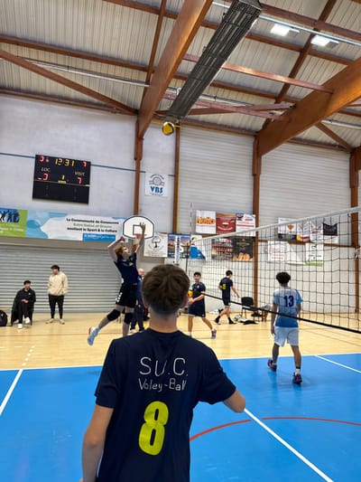 An indoor volleyball match is underway in a gymnasium in Sennecey-le-Grand, France. The scene is observed from behind a male player wearing a dark blue t-shirt with "S.U.C. Voley-Ball" printed in white and a yellow number "8" on his back.
In the mid-ground, a male player in a dark blue jersey and shorts, wearing knee pads, is airborne on the left side of the court, with his hands raised, poised to strike a yellow and blue volleyball suspended in the air near the wooden ceiling beams. A black volleyball net spans the width of the blue court, marked with white and red lines. On the opposing side of the net, a player in a light blue and white jersey with the number "13" is visible, along with other players in dark blue jerseys.
To the left, two male individuals are seated on a bench: one in a black hoodie and another in a beige hoodie. Above them, a digital scoreboard displays "12:31," "LOC - 3," and "VISIT - 7." Below the scoreboard, a basketball hoop with a white backboard is present. Various promotional banners are affixed to the light-colored walls, including "MONTEAU LE GRAND VBS Sennecey le Grand," "La Communauté de Communes Entre Saône et Grosne soutient le Sport & la Culture," and a "Netto" supermarket advertisement. The gymnasium features a high ceiling with exposed wooden trusses, white corrugated metal roofing, and multiple fluorescent lighting fixtures. Large roll-up doors are visible on the right side of the background wall.