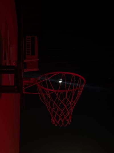 The image captures a dimly lit scene, primarily dominated by dark tones and striking red objects. In the foreground, positioned towards the right, is a miniature red basketball hoop with a net. The hoop appears to be made of metal or rigid plastic, forming a classic basket shape with a loosely woven, net-like structure. Visible at its base are small red springs, suggesting it might be a functional toy or model with a spring-loaded return mechanism.
To the left of the basketball hoop, partially obscured in the darkness, sits another red object that resembles a miniature bleacher or a section of stadium seating. Both red objects rest on a dark, flat surface, possibly a table or a model court, which itself is dimly lit with a reddish hue reflecting from the objects.
The background is almost entirely black, creating a sense of depth and isolating the illuminated objects. A thin, vertical beam of bright white light cuts through the darkness, passing through the center of the basketball net and highlighting a small, spherical white object nestled within the net. This light source creates a dramatic focal point.
There is no discernible activity taking place; the scene appears to be a still life arrangement of miniature sports equipment. The overall impression is one of artificial lighting in a dark environment, possibly indoors. No specific time of day, weather, or explicit location cues are visible within the image itself, beyond the dark, controlled lighting setup. There is no visible text. The location context provided (Ptichnoye, Russia) is not visually represented.