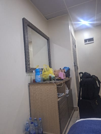 This image depicts the interior of a room located in the city of Close, Nigeria. The specific building or address within Close is not discernible from the visual information.
The room features light-colored walls and a white ceiling constructed with grid-patterned tiles. Artificial overhead lighting illuminates the scene, evidenced by a prominent lens flare in the upper right quadrant of the image.
A wooden dresser or cabinet, comprising a light wood-grained upper section and darker, horizontally striped lower cabinet doors, is positioned against the left wall. A large rectangular mirror with a dark, textured frame is mounted horizontally above the dresser. The dresser's top surface is occupied by multiple objects: several plastic bottles, including those containing blue liquid and clear water bottles, a yellow plastic bag, a small brown bottle, a pink cylindrical container, and tangled white cables. An opaque white plate is visible on an open shelf within the dresser. On the floor to the left of the dresser, five plastic water bottles, some appearing empty, are present.
To the right of the dresser, two black pieces of luggage are stacked vertically on a light-colored tiled floor. The upper luggage piece is a wheeled suitcase with a partially extended handle. Further to the right, a dark door with a visible handle is situated on the wall, and above it, a white rectangular unit is mounted. A blue fabric item, possibly a blanket or towel, is partially visible in the bottom right foreground, suggesting the presence of a bed.