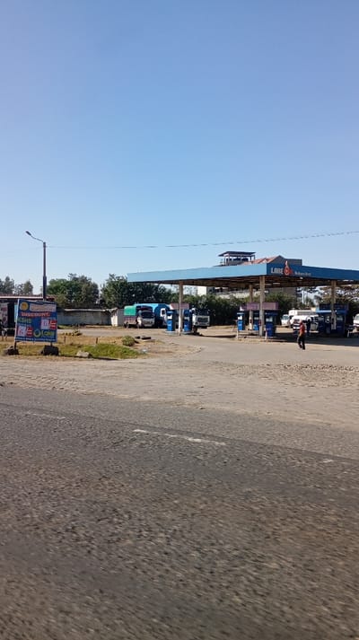 The image displays the Lake Oil gas station situated in Nakuru East ward, Kenya, under a clear blue sky. In the foreground, a dark asphalt road surface exhibits texture and faint white lane markings. To the left, a tall streetlamp pole is positioned next to a blue and yellow billboard advertising "DISCOUNT KSHS SUPER DIESEL" from "LAKE OIL EXPRESS."
The central feature is a light blue-canopied Lake Oil gas station, bearing the inscription "LAKE OIL Nakuru East" on its facade. Multiple blue and white fuel pumps are visible beneath the canopy. Several vehicles are present within the station: a blue truck with a green tarp-covered bed, a white truck, and a white semi-truck cab are positioned at or near the fuel pumps. Additional smaller vehicles are partially discernible further back.
A single individual, wearing a red shirt and dark trousers, walks from left to right across the paved area adjacent to the fuel pumps. Another person is indistinctly visible near the blue truck. The background reveals green trees and bushes, a low concrete wall, and an elevated building structure behind the gas station canopy. The scene is illuminated by bright daylight.