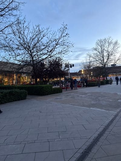 This image captures an outdoor public plaza within the Parquesur shopping center in Leganés, Spain, during twilight. The foreground consists of grey rectangular paving stones with a linear metal drainage grate positioned along the right edge. Dominant bare-branched deciduous trees frame the left and right sides of the scene, with dense green hedges forming planter boundaries in the mid-ground.

To the left, a large building with a multi-story glass facade displays warm interior lighting. In the mid-ground, approximately two dozen individuals are dispersed across the plaza, either standing in groups or walking. They are casually dressed. Several red and white structures, potentially play equipment or decorative elements, are visible among the people. The background features additional buildings, including one with a tiled roof and a partially visible "KFC" sign on the right. Artificial streetlights are illuminated throughout the area. The sky exhibits a light blue and grey gradient with subtle cloud formations, indicative of dusk or early evening.