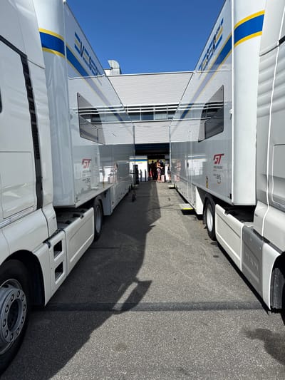 The image shows two white tractor-trailer trucks parked close together on a gray asphalt surface. A few people are visible in the narrow space between the trailers. Blue sky is visible at the upper left. The trailers feature branding for GT World Challenge Europe SRO Motorsports. The scene is located in Campagnano di Roma, Italy.