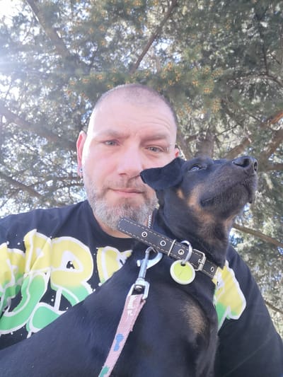 A man with a shaved head, grey beard, and facial piercings is holding a black dog with a leather collar and pink leash in front of a tree. The man is wearing a black t-shirt with yellow and green graphics. The photo appears to have been taken outside, under a tree. Location is potentially Pardubice, Czechia.