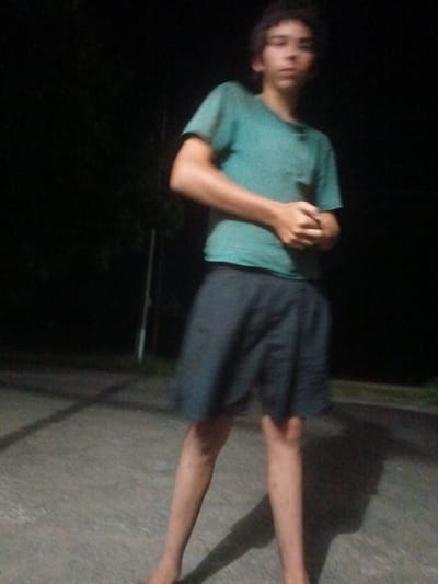 A full-body, slightly blurred, low-light, outdoor photograph shows a young person standing in a driveway or paved area at night. The person is wearing a teal t-shirt and dark shorts. Their hands are clasped in front of their body. A dark, shadowy background suggests trees or foliage. There is no discernible indication of Leontyevskoye, Russia.