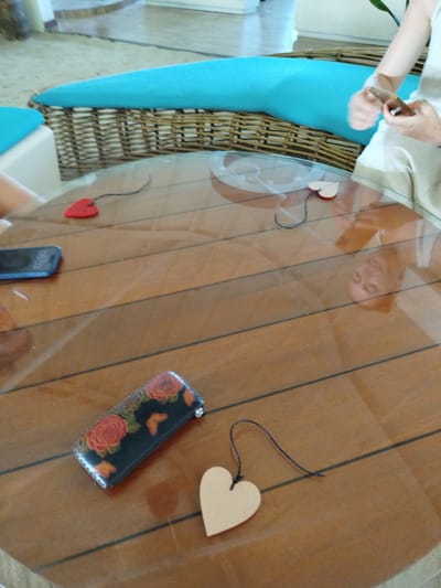 The image shows a clear glass table with a wooden base. On the table, there are several items: a red heart-shaped charm, a light-colored heart-shaped charm, a black rectangular object with a floral pattern, and a blue smartphone. A person's hand is visible on the right side of the image, holding a smartphone. The background shows a wicker chair with blue cushions, suggesting an outdoor or semi-outdoor setting. The scene appears to be located in Gulhi Falhu, Maldives, based on the context provided. The lighting suggests it is daytime.