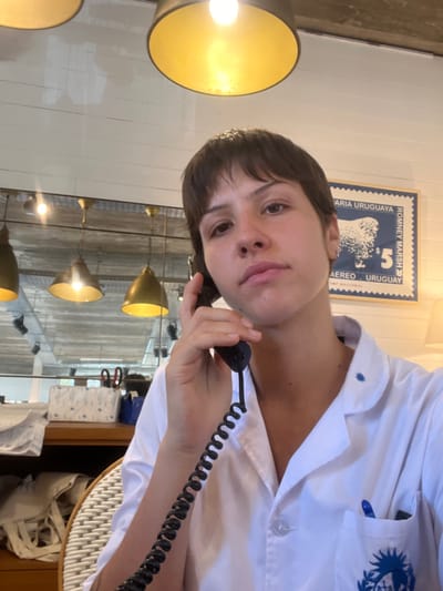 A person holds a black telephone receiver to their ear, their face angled towards the camera. They are wearing a white collared shirt. Behind them is a wall decorated with a framed stamp and a mirror reflecting multiple pendant lights. White walls and a glimpse of a wicker chair are also visible. The lighting is warm, highlighting the lamps and creating a reflective environment.