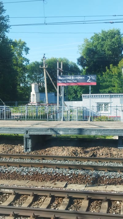 A photograph taken from a train window shows a railway station in Vyshny Volochyok, Russia. A sign at the station reads "Вышний Волочек" (Vyshny Volochek) and "Путь 2" (Track 2). The station appears to be a simple structure with a platform and a small building. The surrounding environment is rural, with trees and vegetation visible. The sky is clear and blue. The photograph is taken in daylight, with sunlight illuminating the scene. The train tracks are in the foreground, with ballast and sleepers visible. The overall impression is one of a quiet, rural railway station.