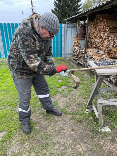 The image depicts an individual outdoors, possibly in a yard or garden, near a structure that appears to be a woodshed. The person is wearing a camouflage jacket, grey pants with reflective stripes, a grey knit hat, glasses, and red gloves. They are crouched down, holding a gardening tool, possibly cleaning or inspecting it. The background shows a wooden structure filled with stacked firewood, suggesting an area where wood is stored for heating or other purposes. There is also a blue and silver corrugated metal fence and a wooden workbench. The ground is a mixture of dirt and grass. The location may be in Talmenka, Russia, based on the instructions provided, but the image itself offers no location-specific information.