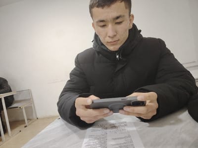 The scene is an interior room located in Kungrad, Uzbekistan. A young male, appearing to be of Central Asian descent, is seated at a light-colored desk in the foreground. He has short, dark hair and is wearing a black puffer jacket. He holds a grey smartphone horizontally with both hands, directing his gaze downwards at the device's screen. A circular grey accessory is attached to the rear of the smartphone. A sheet of white paper with printed text is visible on the desk surface directly beneath the smartphone. In the left background, another individual, wearing a dark jacket, is partially visible, seated at a similar desk with a chair featuring a light-colored seat and a metal frame. The room's walls are plain and light-colored, showing minor surface irregularities. The area is uniformly and brightly illuminated.