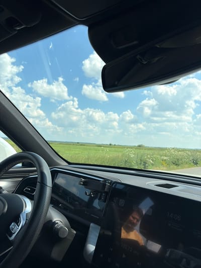 The image is taken from inside a car, looking out through the windshield and side window. The scene outside is a bright, sunny day with a clear blue sky filled with fluffy white clouds. A vast, green field stretches to the horizon, dotted with white flowers, suggesting a rural or agricultural setting.
Inside the car, the dashboard and steering wheel are visible. A large infotainment screen displays the time as 14:06, indicating it's early afternoon. The screen also shows "FM 105.70 MHz," suggesting the radio is on. A reflection on the screen shows the face of a person, likely the driver, looking at the screen. Other details visible on the screen include icons and some text in a Cyrillic alphabet. The steering wheel is black with stitching details.
The overall impression is that of a pleasant drive through the countryside on a summer afternoon. The weather is good, and the setting appears to be Klisheva, Russia, given the location context.