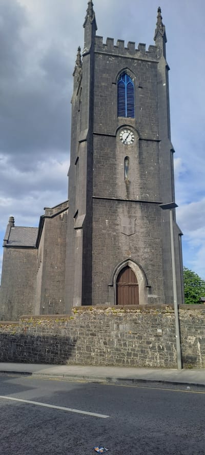 An exterior, low-angle, vertical, full shot captures the imposing stone tower of St. Patrick's Church in Castlebar, Ireland, under a partly cloudy sky. The tower, constructed of dark, weathered stone blocks, features a distinctive arched blue window near its apex. Below the window, a clock face with black Roman numerals on a white background is visible. A small, narrow niche containing a figure is situated below the clock. A smaller, attached section of the church, also made of stone, is visible to the left, with a dark slate roof. The tower and church are bordered by a low stone wall, with sparse green vegetation growing along its top. In front of the wall is a grey paved sidewalk, and further forward, a tarmac road with yellow and white markings. A lamppost stands to the right of the tower. A piece of litter, possibly a candy wrapper, is visible on the road surface in the foreground. No people are present in the scene.