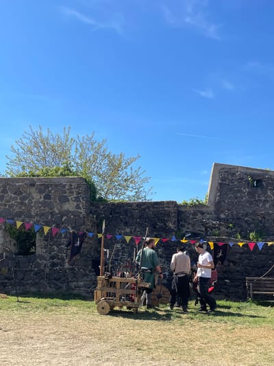 The image shows a section of a stone wall decorated with colorful triangular flags. The wall appears to be part of a larger structure, possibly a castle or fortress. A tree with budding leaves grows near the wall. Three people are positioned near the wall, appearing to be involved in a historical reenactment or display. One person wears a white shirt and dark pants, another wears a light-colored shirt and dark pants, and the third is dressed in green. They are standing near a wooden cart filled with what appears to be historical weapons and equipment. The background includes a clear blue sky with a contrail from an airplane. The scene likely takes place in Hostalric, Spain, based on the presence of a historic fortress or castle and possible reenactment activity.