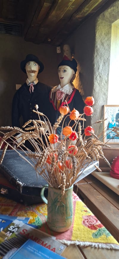 The image depicts a still life arrangement in what appears to be an indoor setting, possibly a museum or a shop in Karja, Estonia. The primary subjects are two mannequins dressed in traditional attire. The mannequin on the left is a male figure wearing a black hat, a black jacket over a white shirt with a red and white checkered cravat. He has a prominent brown mustache. The mannequin on the right is a female figure wearing a black hat with a red trim, a white collar with lace, and a richly decorated outfit featuring red, yellow, and black elements. She has large blue eyes and red lips.

In the foreground, a bouquet of dried wheat stalks with orange Chinese lanterns (physalis) is placed in a light green, patterned ceramic jug. The jug sits on a colorful woven mat with a floral design. Behind the jug and to the left, a dark, aged suitcase is visible. In the background, the ceiling is constructed of rough-hewn wooden beams. The walls appear to be made of rough, textured material, possibly plaster or stone. To the far right, a framed picture or artwork is partially visible, depicting a scene with blue hues. Several travel brochures or books with blue covers are laid out on the mat in the lower left corner of the image, one of which prominently displays the word "SAAREMAA" in red letters. The lighting suggests an indoor environment, with natural light possibly coming from an unseen window. The overall impression is one of cultural display and perhaps a nod to harvest traditions.