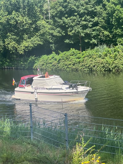 A motorboat named "Seebar" is navigating a body of water, likely a canal or river, near the bank lined with lush green trees and vegetation. The boat is white with a red canopy and has flags on its stern. A person is visible sitting on the upper deck of the boat, looking to the right. A metal fence runs along the foreground, separating the water from a grassy area with some yellow wildflowers. The scene appears to be set in Teltow, Germany, given the context provided. The water is rippled, indicating movement, and the sunlight suggests it is daytime.