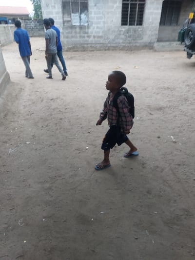 The scene depicts an outdoor setting in Mwandege, Tanzania, on a dirt path. In the foreground, a young child, appearing to be a toddler or preschooler, walks from right to left, carrying a black backpack. The child is dressed in a red, white, and dark blue plaid long-sleeved shirt, dark shorts, and light blue flip-flops. Their head is turned slightly right, looking upwards. In the background, to the left, three adult males are walking away from the camera, moving from left to right. The leftmost male wears a light blue shirt and light grey pants. The central male wears a grey t-shirt and dark grey pants. The rightmost male wears a blue t-shirt and dark pants. Behind them, a grey brick building with barred windows is visible, along with a partial view of a vehicle with a rear-mounted spare tire on the far right. The ground is unpaved dirt.