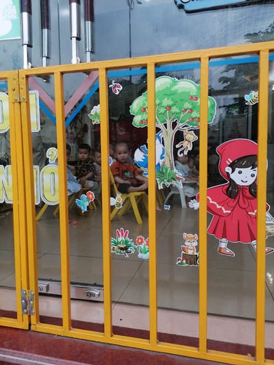 This image offers a glimpse into a kindergarten or daycare classroom in Ha Tu Ward, Vietnam, seen from outside through a bright yellow metal gate. The gate, composed of vertical bars and horizontal rails, occupies the immediate foreground, with a hinge visible on its left side.
Inside the room, several young children, likely toddlers or preschoolers of Asian descent, are seated on small yellow chairs around low tables. One child in a black patterned shirt and another in an orange shirt both look directly towards the viewer with calm, neutral expressions. Other children are partially visible behind them or obscured by the gate's bars.
The large glass window that forms the wall of the room is extensively decorated with colorful, cartoon-style cutouts. These decorations include a large green tree with red apples and a monkey, a character resembling Little Red Riding Hood in a red cape, a blue fox, a squirrel on a tree stump, a bee, a ladybug, a frog, and various floral and butterfly accents. Large yellow letters, spelling out "N10," are also affixed to the left side of the window.
The children appear to be engaged in a quiet activity, such as a lesson or snack time, given their seated positions. The room's floor is tiled, and two silver lock mechanisms are visible on the bottom frame of the glass door. Reflections in the upper portion of the window suggest it is daytime, capturing glimpses of external structures or the sky. The vibrant decorations and miniature furniture clearly identify the setting as a childcare facility.