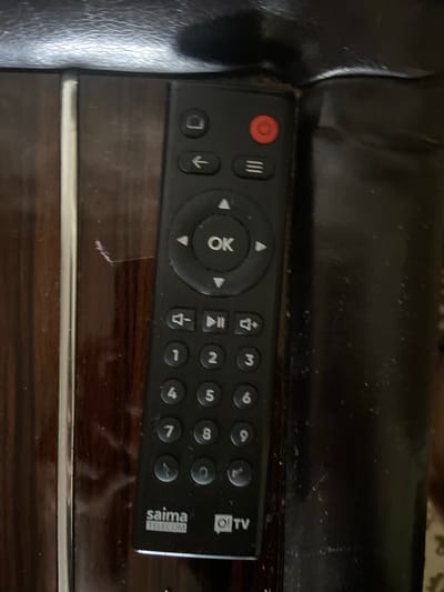 The image shows a black TV remote control with white buttons and text. The remote has numbered buttons from 0 to 9, as well as navigation buttons and a red power button. The brand name "saima TELECOM" and "Oi TV" are visible at the bottom of the remote. The remote is resting on a dark, wooden surface that appears to be a table or furniture. The lighting in the image is dim, and the surface has some dust and scratches. The background is out of focus, but it appears to be a dark, possibly upholstered surface. There is no indication of the city of Kojomkul, Kyrgyzstan, in the image itself or in the provided crops.