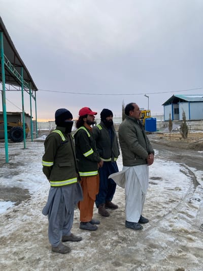 Four adult males stand in a line on a snow-dusted, muddy ground under an overcast sky at a construction or temporary facility site in Kan Mehtarzai, Pakistan. The men are dressed in warm clothing, including jackets, shalwar trousers, and various head coverings. From left to right:
1.  A man wearing a dark beanie, a dark face covering, a dark green jacket with reflective yellow stripes, and gray shalwar. His hands are behind his back.
2.  A man with long dark hair and a beard, wearing a red baseball cap, a dark green jacket with reflective yellow stripes, and brown shalwar. His hands are behind his back.
3.  A man with a dark beard, wearing a dark beanie, a dark puffer jacket, and dark blue shalwar. His hands are clasped in front.
4.  A man with short dark hair, wearing an olive green puffer jacket and white shalwar kameez. His hands are clasped in front.

The men are facing towards the right side of the frame, appearing to observe something outside the image. In the background, to the left, is a metal-framed shelter with a light roof, partially obscuring a yellow heavy machinery vehicle. To the right, beyond a fence, several modular, pre-fabricated buildings with light blue bases and white tops are visible, some with snow on their roofs. A blue cylindrical tank and several small trees are also present in the middle ground, beneath a light pole. The ground is a mix of dirt, melted snow, and ice, with visible tracks.