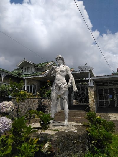 An outdoor, daytime image captured in Nathene, Laos, features a weathered, light-colored female statue positioned centrally in the foreground. The statue, standing on a concrete base, is partially draped with fabric around its lower body, holding a spherical object in its right hand near the chest and a draped cloth in its left hand. Behind the statue, a multi-level building with a distinctive green-tiled roof, white walls, and a stone foundation is visible. The building exhibits multiple windows, a porch with white railings, and a dark satellite dish mounted on the roof structure to the right. The immediate foreground is populated with various green plants, including clusters of light purple/white hydrangeas on the left. The upper portion of the scene shows a sky with significant white cumulus cloud cover and patches of clear blue sky. A dark utility line crosses the sky diagonally from the upper left. No human figures are present.