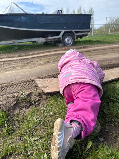 A child lies prone on the ground in Тоншалово, Russia, near a dirt road and a parked trailer. The child wears a pink and white striped jacket, pink pants, striped socks, and light-colored shoes with a velcro strap. The ground is a mix of dirt and green grass, and a piece of plywood lies between the child and the dirt road. The trailer, attached to a vehicle, is visible in the background on the left side of the image. The weather appears to be clear and sunny.