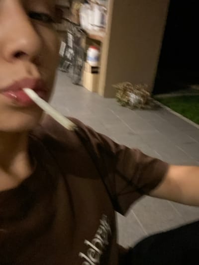 A close-up, blurred, angled image shows a person with a pale tongue sticking out, holding a white object (possibly a cigarette) between their lips. They are wearing a brown shirt. In the background are elements of a brick building, sparse vegetation, and what appears to be a cluttered outdoor space with boxes or materials, all under dim lighting. The image is low quality and lacks sharpness.