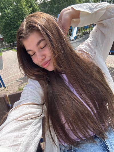 The image captures a young woman from an angled perspective, seemingly leaning back or positioned with her head tilted. Her eyes are closed or nearly closed, and her lips are softly parted, indicating a relaxed and tranquil expression. She has long, straight, medium-brown hair that cascades over her chest and shoulders.

She is dressed in a light-colored, possibly off-white, long-sleeved shirt made of a subtly textured or crinkled fabric, with a visible buttoned cuff on her left arm. A hint of a light purple garment is visible beneath her hair, along with blue denim jeans.

The scene is outdoors on a bright, sunny day, with ample natural light creating highlights on her hair and clothing. In the background, lush green trees are visible to her left. To her right, there is a paved area with light-colored brick or concrete blocks, suggesting a park, square, or recreational space in Речицы, Russia. Glimpses of blue and darker structures, possibly playground equipment or benches, can be seen behind her. There is no visible text in the image.