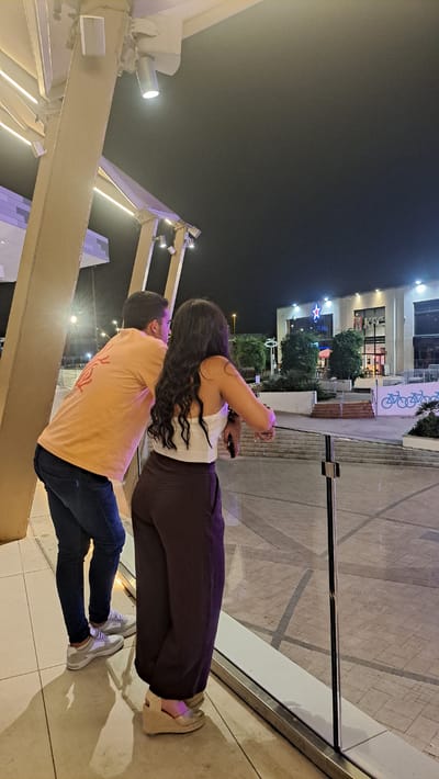 The image shows two people, a man and a woman, standing outdoors at night. The woman is wearing a white crop top and dark trousers, while the man is wearing an orange t-shirt and blue jeans. They are standing close together, with the woman appearing to lean into the man. The scene is lit by artificial lights, suggesting it is evening or night.
In the background, there is a modern building complex with several trees. One building has signage that is partially visible, including what appears to be a star logo and letters that might spell "STAR". There are also lights on the buildings and string lights visible in the foreground. The ground is paved with geometric patterns. The overall impression is that of a public area, possibly a shopping center or a plaza, in a city. The lighting suggests a pleasant evening, and the proximity of the two individuals hints at a relaxed or romantic interaction. No specific weather conditions are discernible from the image. The location context of Alcorcón, Spain, is provided separately and not visible within the image itself.