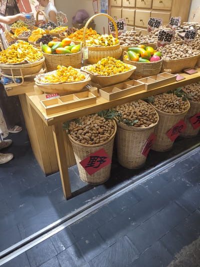 This is a medium shot of a market stall featuring a variety of dried fruits and possibly tamarinds, located in Wuhou District, China. The stall is constructed of light-colored wood and displays goods in woven baskets and wooden bowls. Prominently featured are several large baskets filled with tamarinds, some with red paper tags indicating pricing or product information. To the left, there are baskets overflowing with yellow, rolled dried fruit. In the center, a basket contains colorful mangoes. Above the stall, wooden drawers are visible, and various signs with Chinese characters are displayed. In the foreground, a woman's legs and feet are visible, wearing casual footwear, suggesting she is a customer browsing the products. The floor is dark gray slate tiles. The overall scene depicts a vibrant market environment with a focus on fresh produce and dried goods.