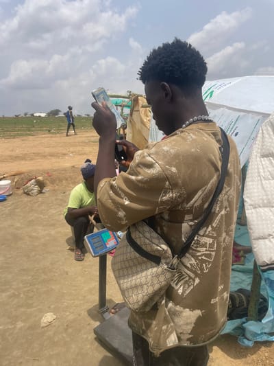 An African male with dark hair is counting Nigerian currency in an outdoor setting in Maiyanga, Nigeria. He is wearing a brown shirt with a white pattern, a silver chain necklace, and a Gucci crossbody bag. A digital scale is next to him on the ground. In the background, other people are present, and a field is visible. Bags of "DANGOTE POLYPROPYLENE" are piled nearby. The ground is dirt.