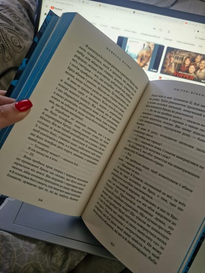 The image displays an open book held by a person's hand with red nail polish. The book's pages are filled with text in Ukrainian. In the background, a laptop screen is visible, displaying a webpage with a title suggesting it is related to YouTube, featuring several thumbnails of videos. The overall scene appears to be indoors, likely in a casual setting like a home. There is no information in the image to suggest it is located in Bratkivtsi, Ukraine.