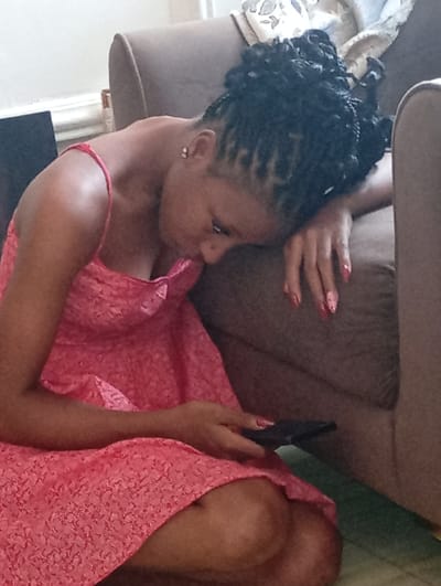 A dark-skinned female with braided hair is seated on the floor next to a brown sofa. She is wearing a light red dress. Her head rests on the sofa cushion, and her right hand is on the cushion. Her left hand holds a black cell phone. The location is potentially a residential setting, but there are no objects or elements in the image to confirm the city of Basawa, Nigeria.