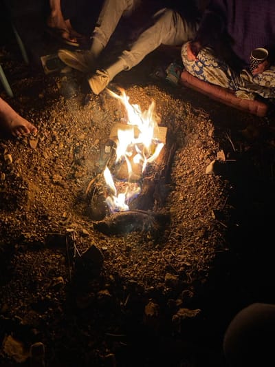 The image displays an outdoor nighttime scene in the city of Mas'ade, Israel, centered around a small, active bonfire. The fire burns within a shallow depression dug into unpaved ground composed of dirt, small rocks, and gravel. Visible elements of the fire include several pieces of wood, some appearing as cut logs, emitting bright orange and yellow flames with blue bases.
Multiple individuals are present, observed through their lower extremities. To the immediate bottom-left, a bare human left foot rests on the rocky ground. Above it, to the left of the fire, a leg encased in light-colored trousers and a light-colored shoe (possibly a sneaker) is visible, with the foot positioned close to the flame. Partially obscured behind this leg is another foot, possibly wearing a sandal. To the right of the fire, the lower body of an individual is partially visible, covered by a blue, white, and brown patterned blanket. This person appears to be seated or reclining on a reddish-brown mat or sleeping pad. A hand, likely belonging to this individual, is holding a white cup adorned with black polka dots. The scene depicts a stationary gathering around the fire.