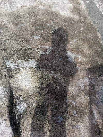 An upright, dark, human-shaped shadow is cast onto a textured ground surface in Cipendawa, Indonesia. The shadow, appearing to be from an individual with arms positioned across or in front of the torso, is distinct, indicating strong, directional light, likely sunlight. The ground surface is mottled with varying shades of gray, brown, and possibly green, consistent with weathered concrete or asphalt, showing signs of dirt, wear, and possibly biological growth. Grooves and irregular patterns are visible across the surface. No other individuals or objects are discernible in the immediate scene.