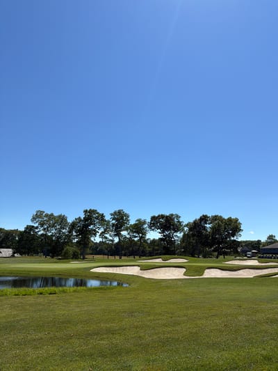 A bright, sunny day on a golf course in the Town of Southampton, United States of America, is captured in this image. The foreground is dominated by lush green grass. In the mid-ground, a calm body of water reflects the sky, bordered by more manicured grass and several sand traps with distinct, undulating edges. Beyond the water and sand traps, a gently sloping green is visible, marked by a flag. A line of mature trees with full, green foliage forms a natural backdrop, separating the golf course from a few buildings in the distance. One building appears to be a clubhouse with a grey roof, and another is a residential structure with a beige facade. Several golf carts are parked near the buildings. The sky is a clear, vibrant blue, with a faint lens flare in the upper right quadrant.