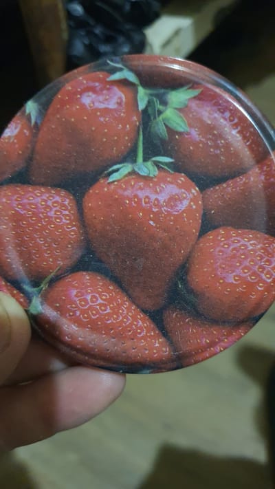The image shows a close-up of a circular object with a print of strawberries on it. The object appears to be a lid or a flat surface with a glossy finish. Several strawberries are depicted in various sizes and arrangements, with green stems and leaves visible on some. The lighting in the image creates reflections on the surface of the strawberries. A person's fingers are visible in the lower left corner, holding the object. The background is blurred, showing what appears to be a dark, cluttered surface, possibly a shelf or table. There is no indication of the city of Karankul, Uzbekistan, in the image or its context.