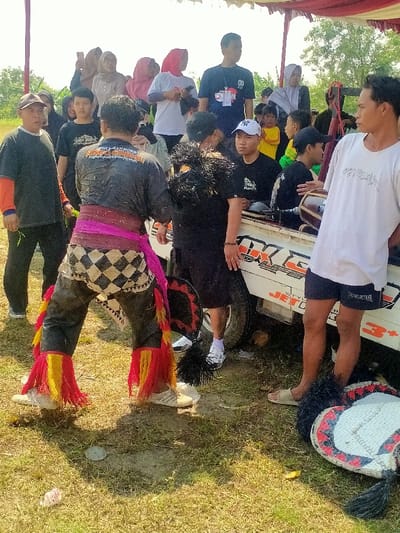 The image depicts an outdoor scene in Celep, Indonesia, under a red and white fabric canopy. In the foreground, a male performer stands with his back mostly to the camera, wearing a dark, possibly wet or muddy, long-sleeved shirt, a wide pink sash around his waist, and checkered pants over dark trousers with prominent red and yellow fringes on the lower legs, standing in light-colored sandals. To the right of the performer, a man in a black t-shirt and dark shorts stands beside a white pickup truck, holding a dark, feathery object. On the far right, another man in a white t-shirt and dark shorts stands near the truck, looking towards the left, holding a kendang drum. Two large, circular, woven props with black feathery fringes and red and white patterns rest on the dry, sparse grass in the lower right. The white pickup truck, visible from the side, has text including "JET C-" and "3+". In the mid-ground and background, approximately fifteen spectators, including men, women (some wearing headscarves), and children, observe the scene from various positions; some individuals are holding mobile phones, and one person is seated in the bed of the pickup truck. The environment is bright and sunny, with foliage visible in the distant background.
