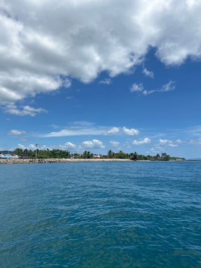 The image shows a coastal scene in Tidore Islands, Indonesia. The foreground is dominated by a vast expanse of blue ocean with gentle ripples on its surface. The middle ground features a rocky shoreline, behind which lies a strip of land densely populated with trees, including palm trees, and various buildings with red and blue roofs. A green flag is visible on a flagpole amidst the foliage, and a street lamp stands prominently. In the distance, the sky is a vibrant blue, adorned with scattered white clouds. The overall impression is a tranquil, tropical coastal environment.