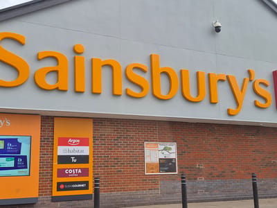 The image shows the exterior of a Sainsbury's supermarket. The large, orange "Sainsbury's" logo is prominently displayed above the entrance on a gray facade. Below the logo, the exterior brick wall features several signs. A brightly colored sign lists various services including "Argos", "Habitat", "Tu", "Costa Coffee", and "Sushi Gourmet." Another sign advertises Sainsbury's branded products, and a third poster displays a chameleon and information. A security camera is visible on the facade above the logo. Black bollards stand in front of the brick wall. The location is in Pity Me, United Kingdom.