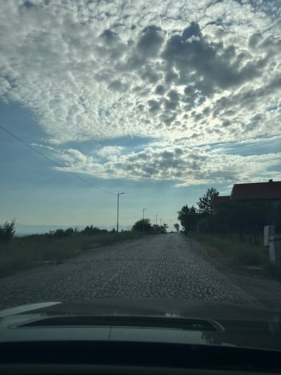 The image captures a view from inside a vehicle, looking out at a cobblestone road leading uphill towards a rural village. The sky is filled with dramatic, scattered clouds, with sunlight breaking through in places, creating a somewhat moody atmosphere. A single telephone wire stretches across the upper portion of the frame. Along the side of the road, there are sparse patches of grass and some trees, with hints of distant hills in the background. A few streetlights are visible along the road, and a couple of houses with red roofs are situated on the right side of the frame. The hood of the vehicle is in the foreground, slightly obscuring the bottom of the image, and a Mercedes-Benz logo is visible on the hood. The scene appears to be located in or near the village of Ivanyane, Bulgaria, based on the context provided.