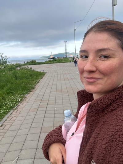 A woman with freckles and brown hair is taking a selfie outdoors in Vilyuchinsk, Russia. She is wearing a pink shirt and a brown fuzzy jacket. She is holding a clear plastic water bottle. In the background, there is a paved walkway, a grassy area with small white flowers, a body of water, a tugboat, and a cloudy sky. A tower with lights is also visible in the distance.